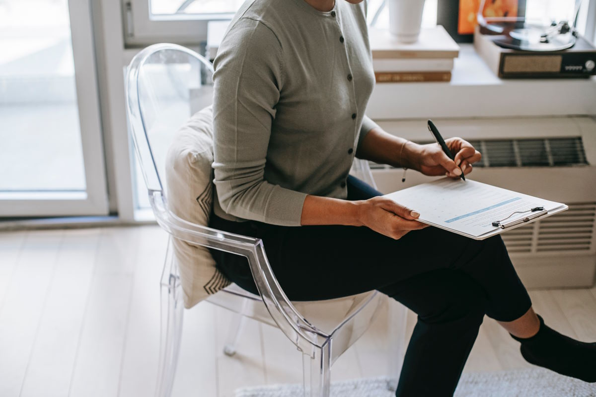 Person sat on a clear plastic chair holding a clipboard and pen