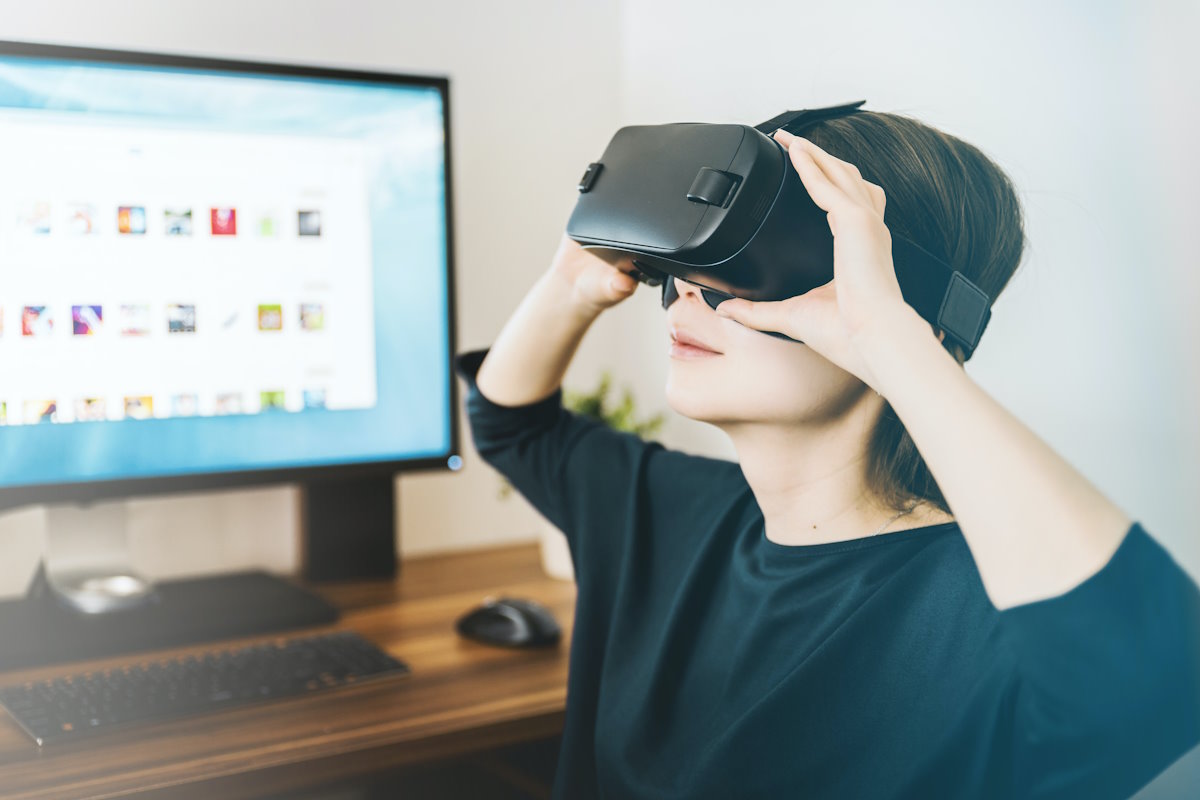 A woman wearing a VR headset, sat in front of a desk with a computer and a potted plant
