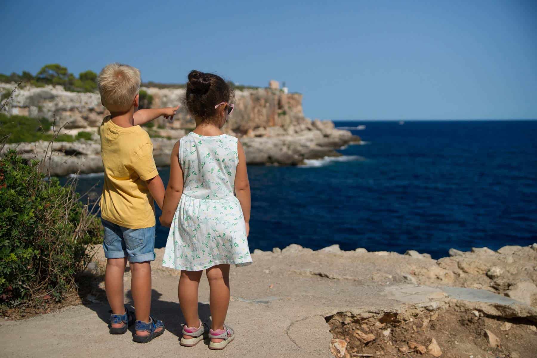 Two children holding hands on a cliff overlooking water, with the child on the left pointing at the horizon